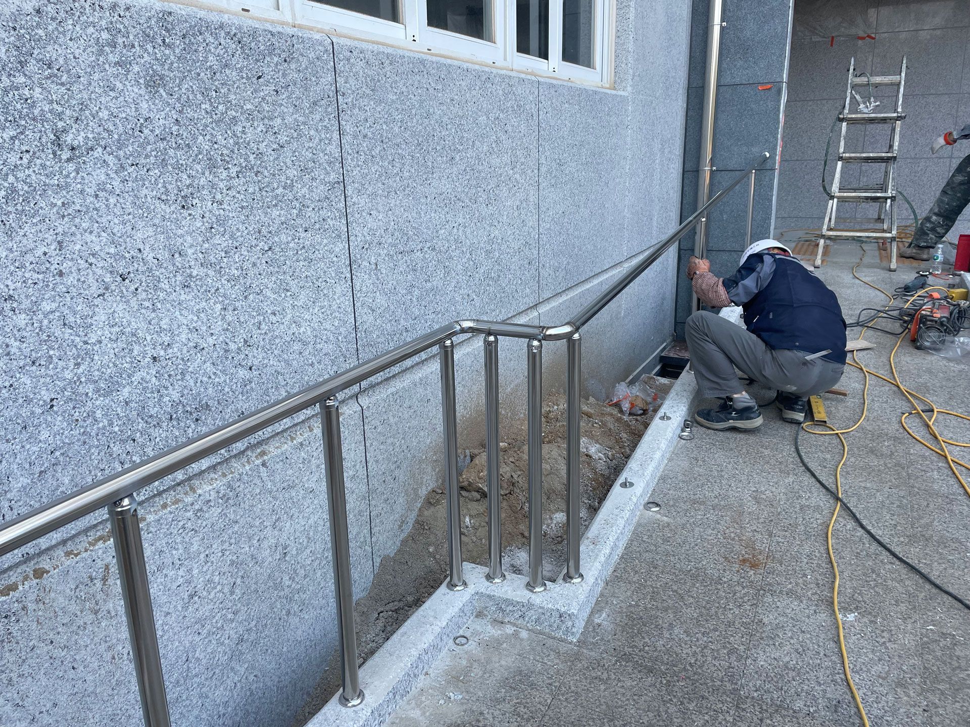 Worker installing a metal handrail along a concrete building exterior, with tools and ladder nearby.