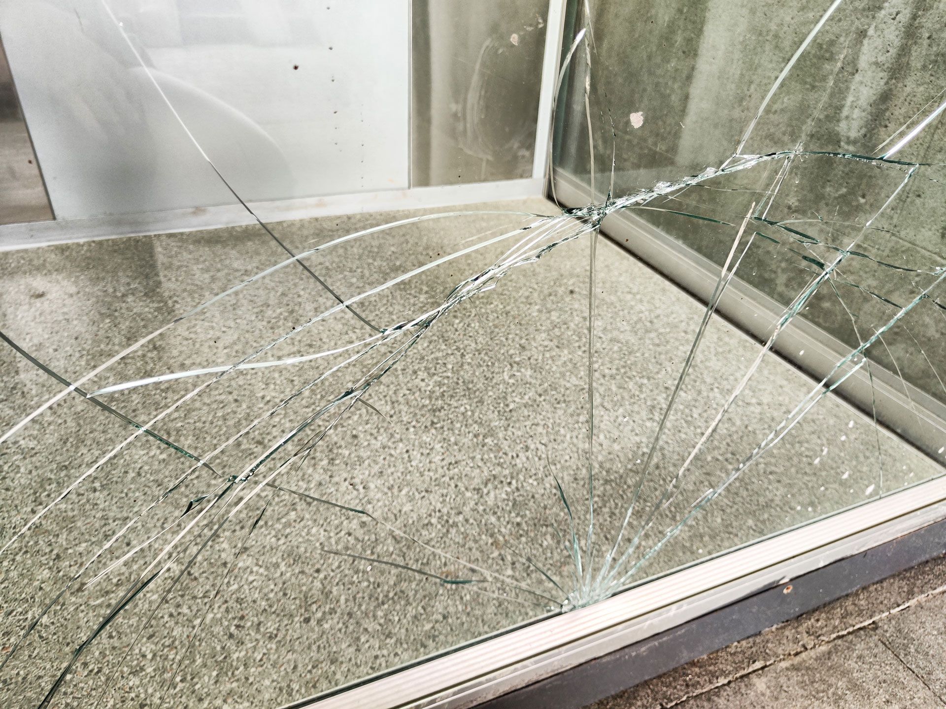 Cracked glass panel with spiderweb fractures on a reflective metal surface near a wall.