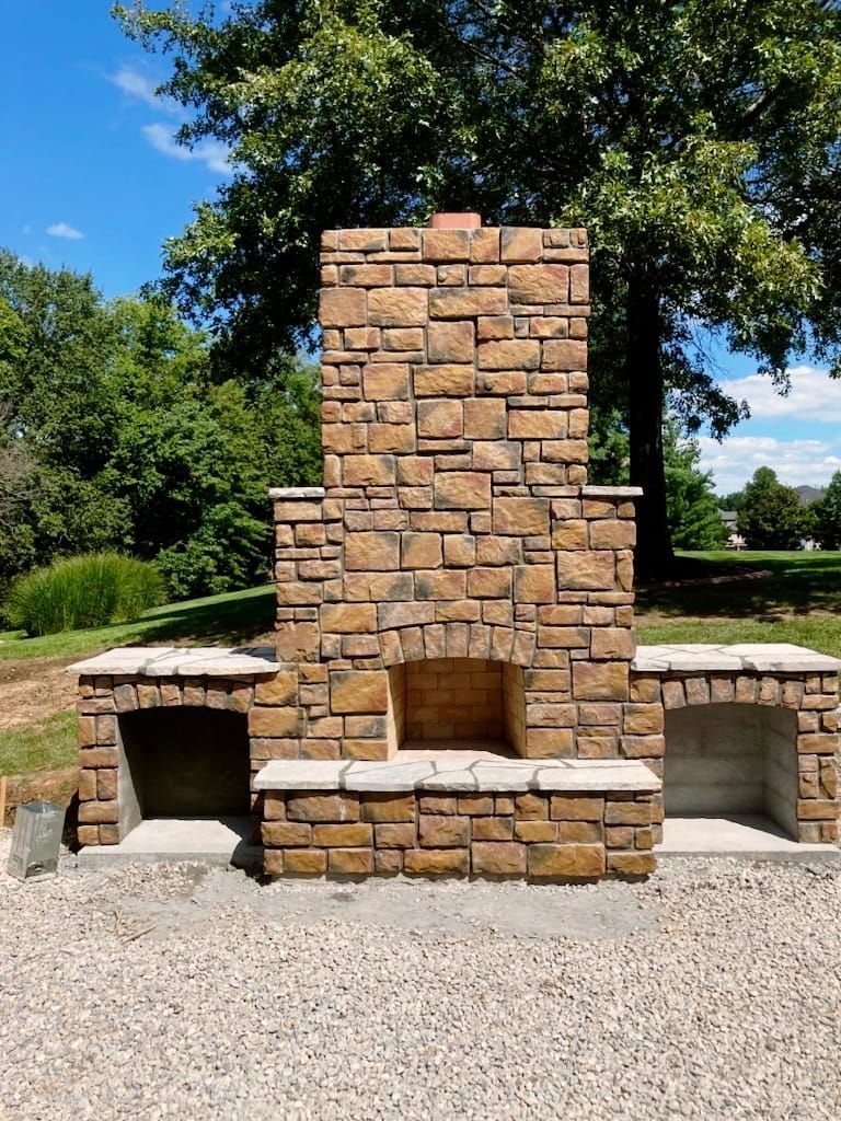 Stone outdoor fireplace with side storage, built in a grassy area with trees and a blue sky.