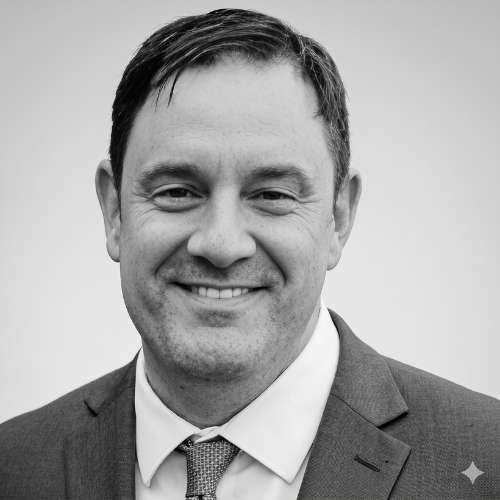 A headshot of a person in a suit and tie smiling, set against a blurred background of a house exterior.