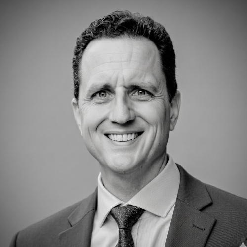 A professional headshot of a smiling person in a suit and tie, captured against a softly blurred background.