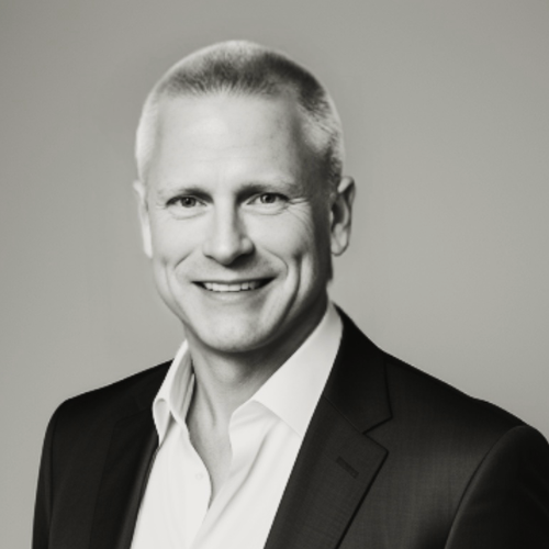 Black and white headshot of a smiling person in a suit jacket and dress shirt against a plain gray background.