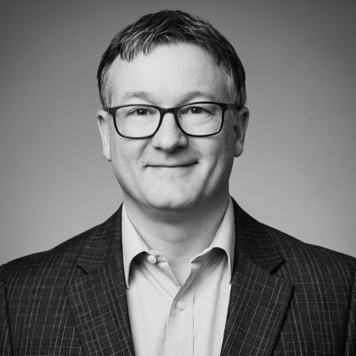 Head-and-shoulders portrait of a person wearing glasses and a plaid blazer, smiling slightly against a blurred background.