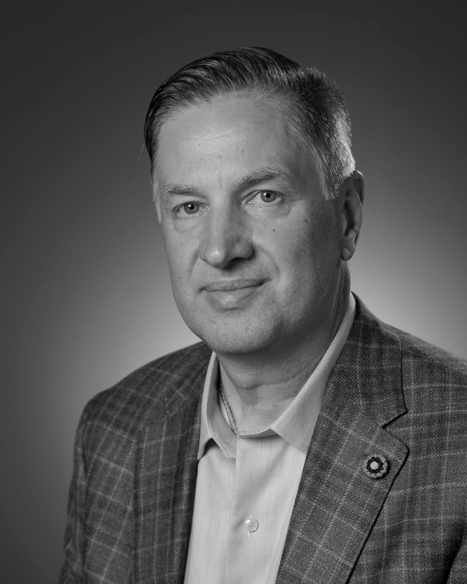 Headshot of a man with short hair wearing a collared shirt and a textured blazer against a solid dark background.