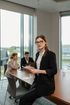 A business professional holds a document while posing in a modern office meeting room with colleagues in the background.