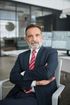A professional with a beard, wearing a suit and red tie, sits with arms crossed in an office with computer screens.