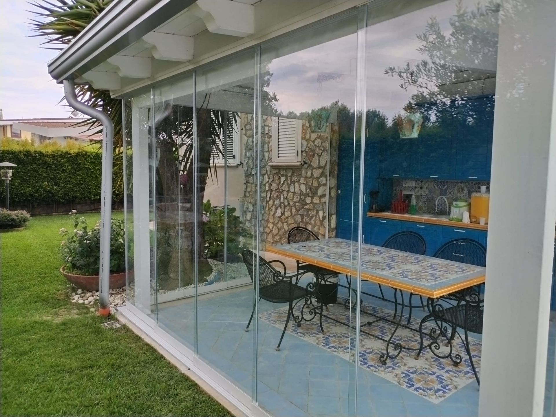 Patio vetrato con tavolo da pranzo, sedie e cuscini colorati accanto a un prato.