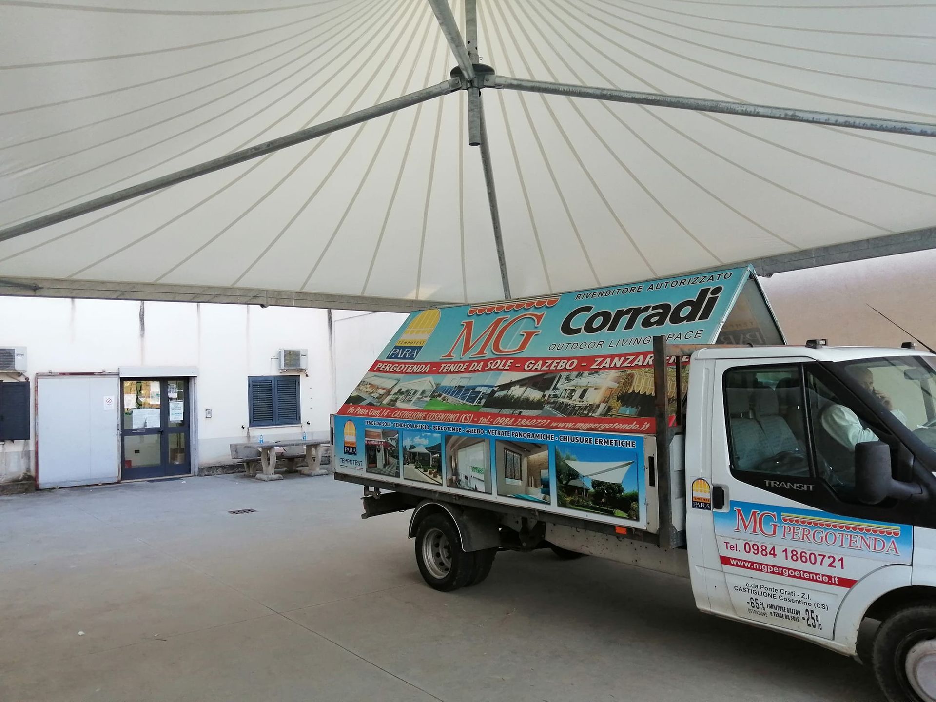 Un camioncino bianco per le consegne con insegne colorate è parcheggiato sotto un tendone in un cortile all'aperto.