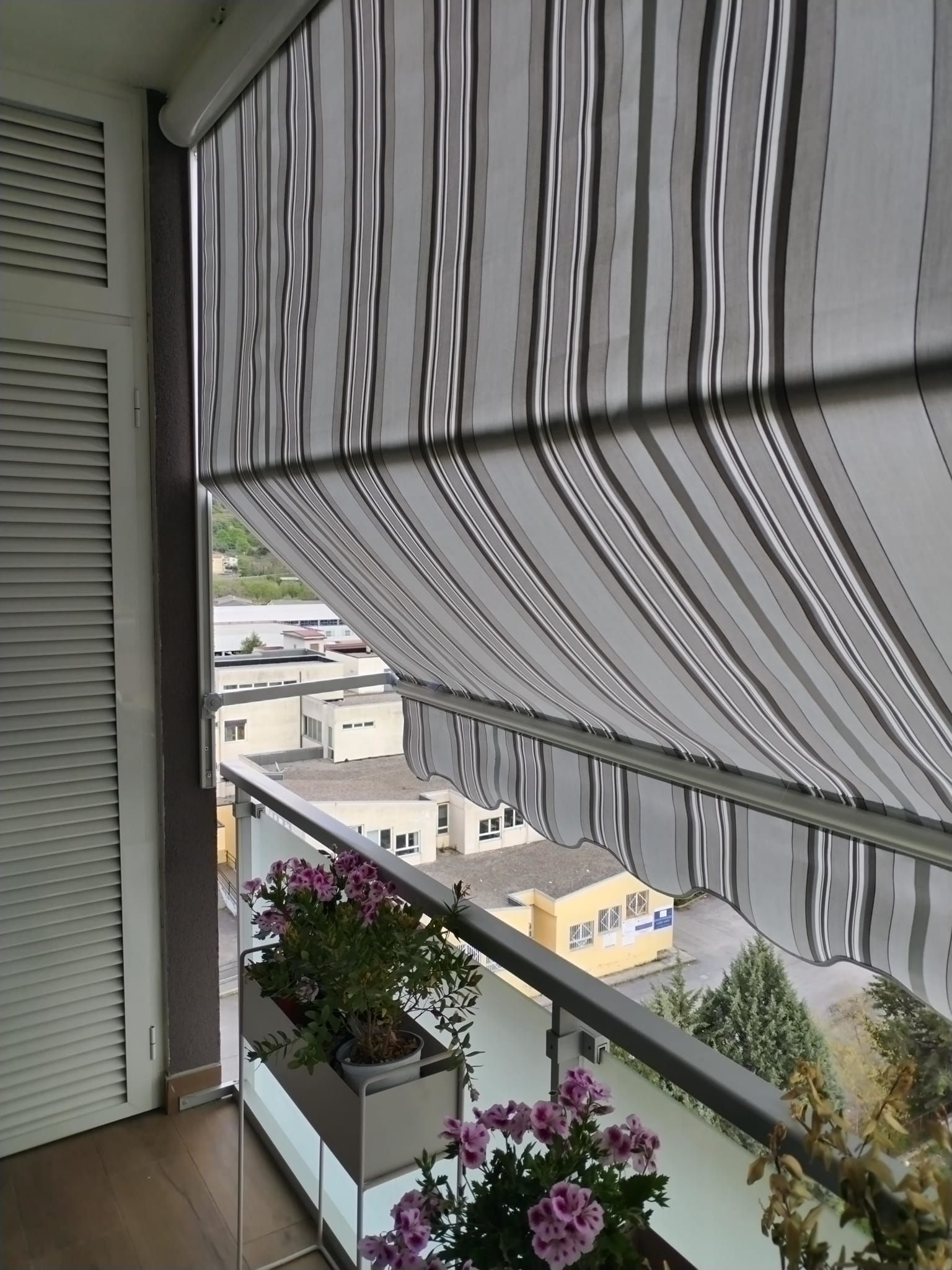 Balcone con tenda da sole a righe, fioriere e vista su una strada della città sottostante.