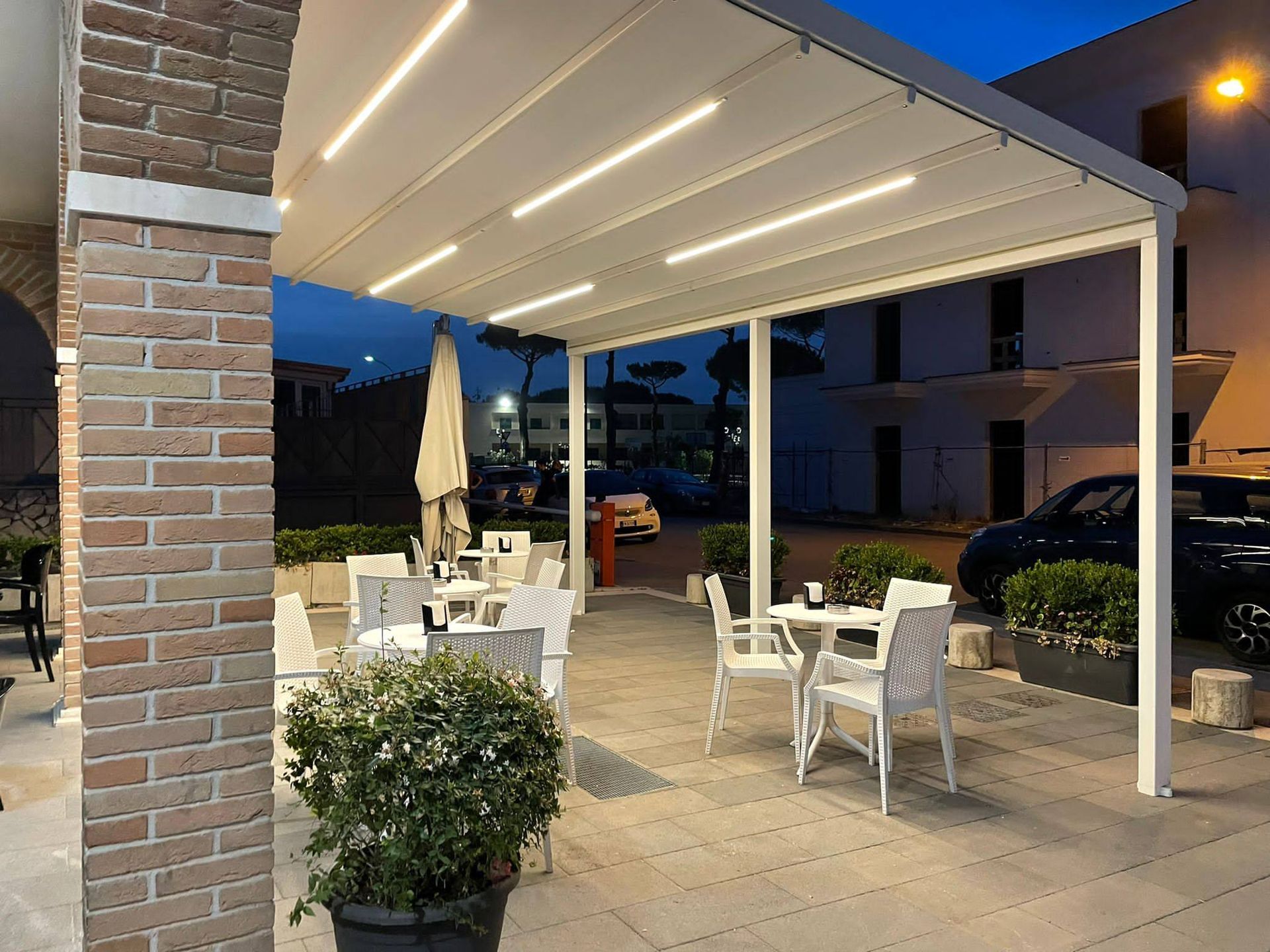 Dehor esterno del caffè con tavoli e sedie bianchi sotto una tenda illuminata al crepuscolo