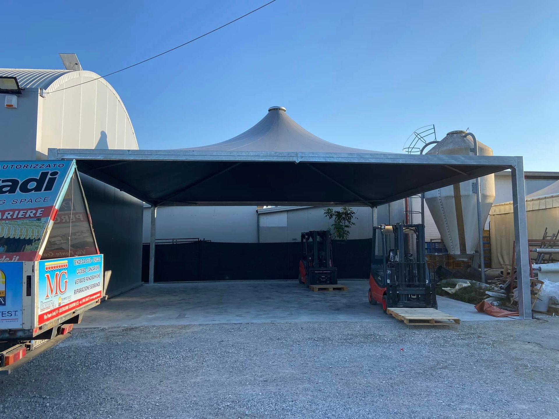 Grande tendone nero su un piazzale ghiaioso con attrezzature e camion sotto un cielo azzurro e limpido.