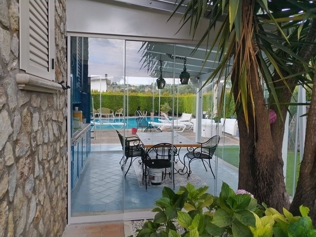 Tavolini da giardino a bordo piscina, con muretto in pietra, ombrelloni bianchi e palme.