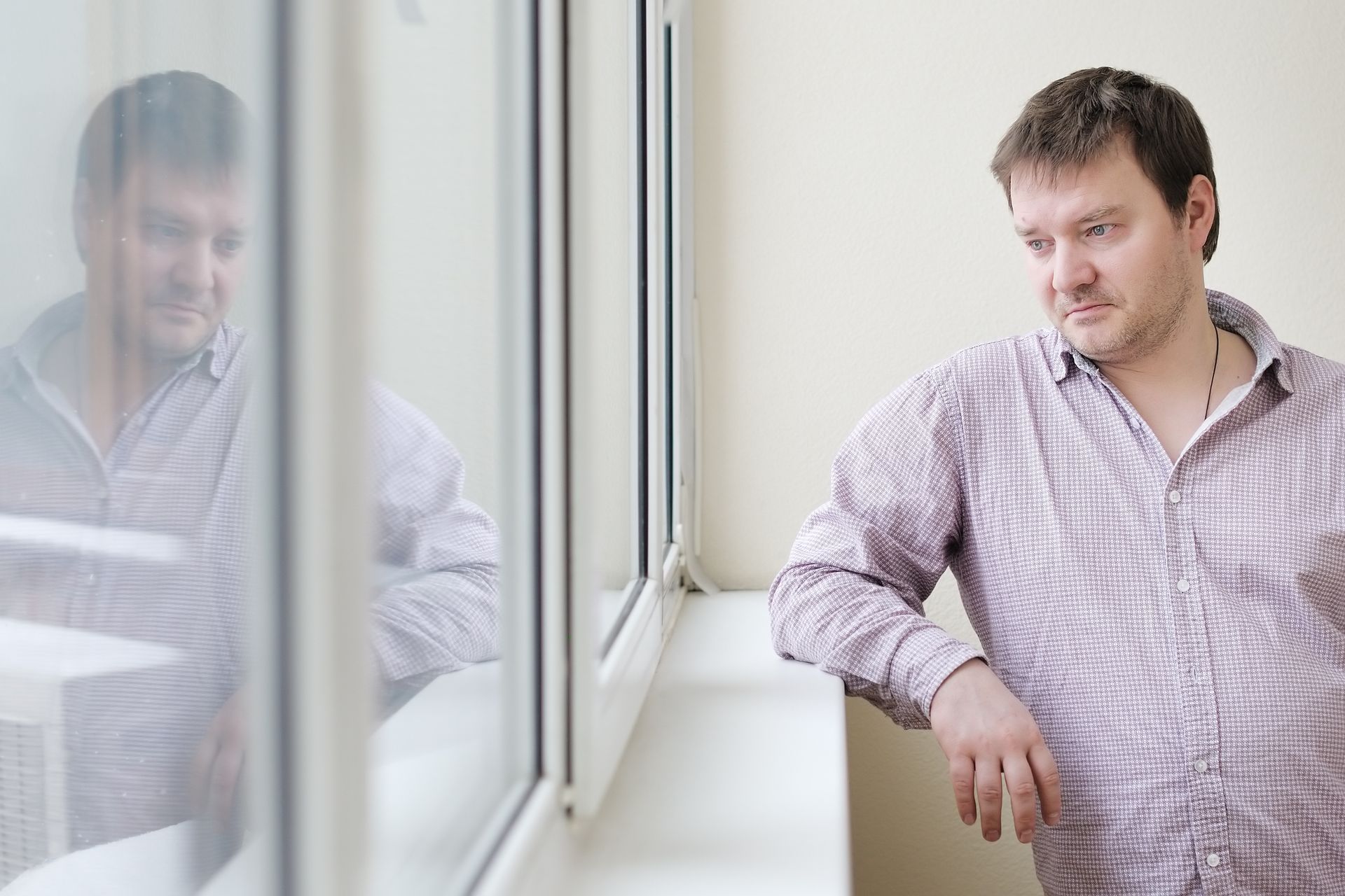 Man standing by a window in a light checkered shirt, looking out with his reflection visible in the glass