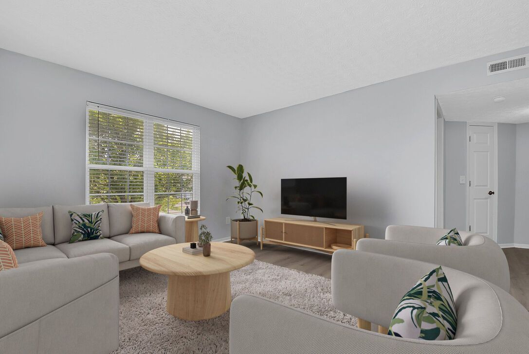 Bright modern living room with beige sofas, round wood coffee table, TV console, and window blinds