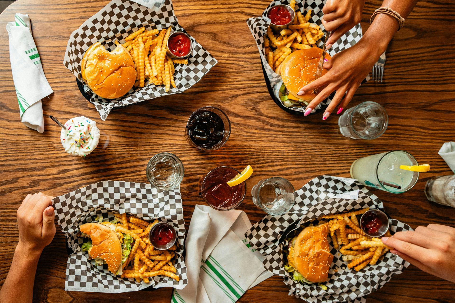 People sharing burgers and fries at a wooden table with checkered paper and drinks