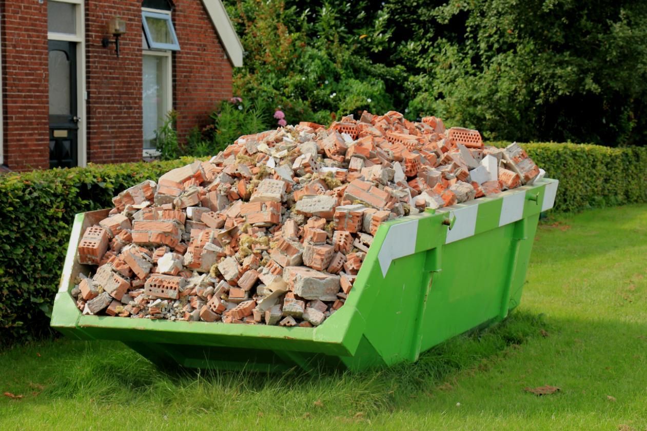A Green Dumpster Filled With Bricks is Sitting in the Grass in Front of a House — Chooks Sand And Gravel In Paget, QLD