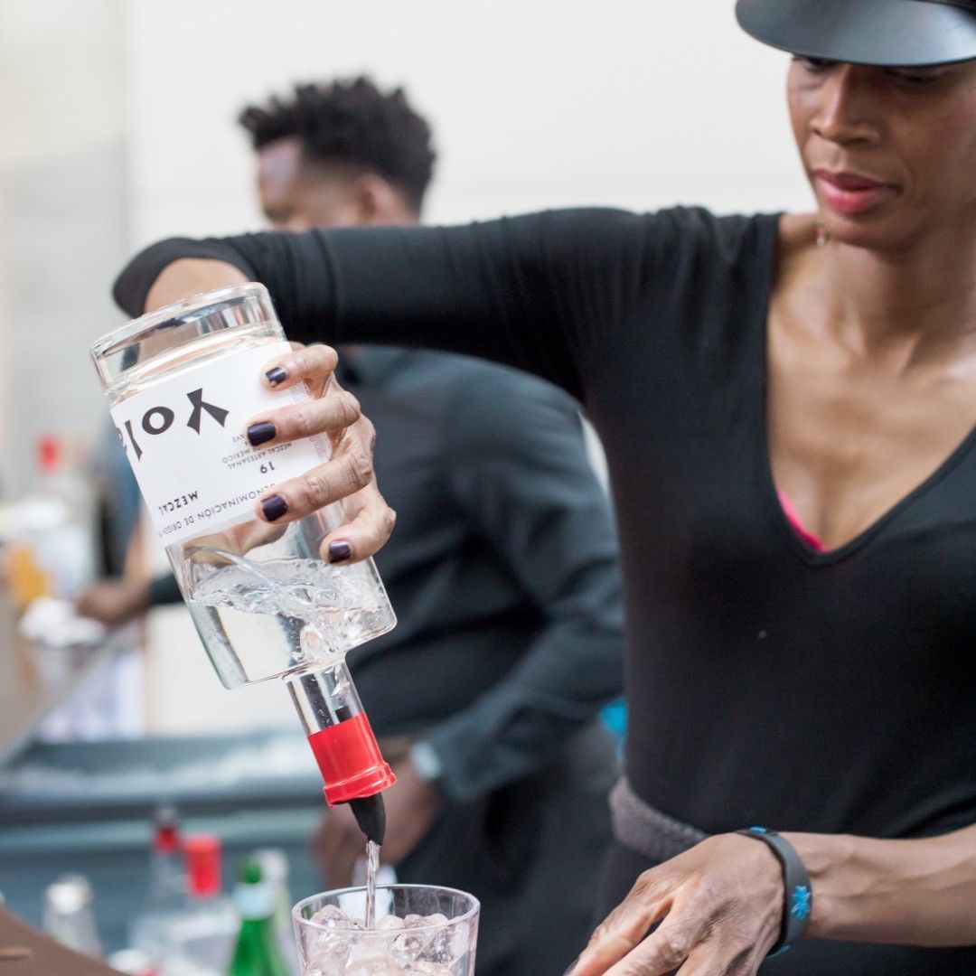 Person pouring clear liquid from a bottle into a glass in a bar or kitchen setting