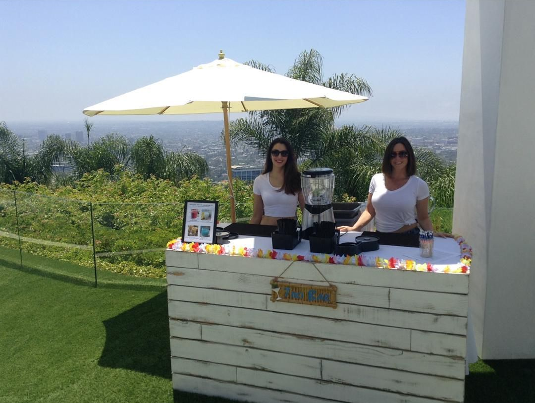 Two vendors at an outdoor drink stand under a white umbrella, with greenery and ocean view behind them.