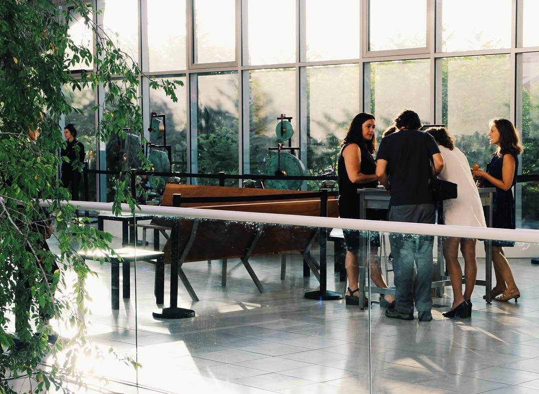 People gathered around a grand piano in a bright glass-walled lobby with plants.