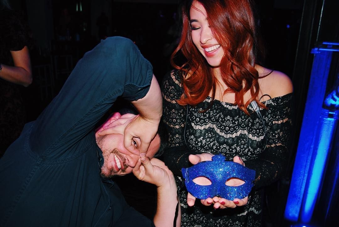 Two people at a nightclub, smiling; one holds a blue masquerade mask.
