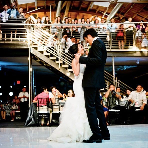 Bride and groom sharing their first dance in a reception hall, guests watching from tables and balcony.