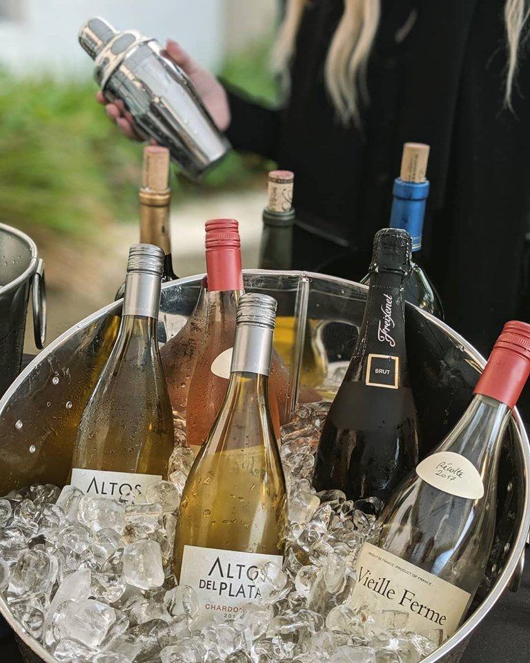 Assorted wine bottles chilling in an ice bucket outdoors, with a hand holding a bottle opener