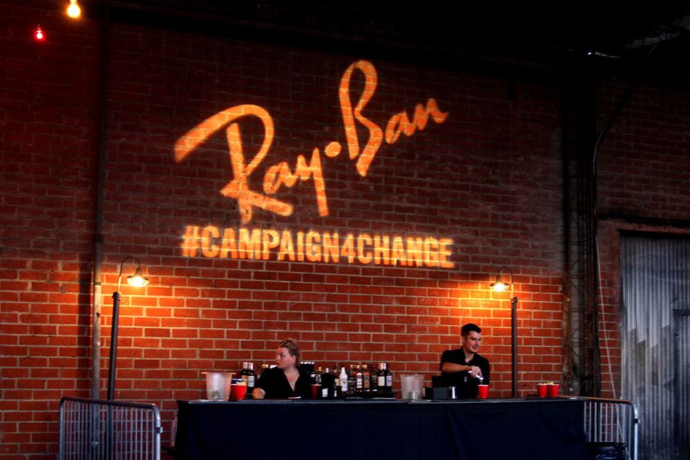 Ray-Ban event stage with brick backdrop, glowing logo, hashtag, and two people behind a long table