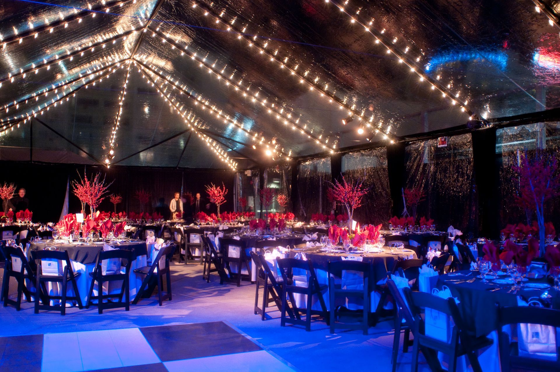 Decorated banquet hall with round tables, red floral centerpieces, and string lights under a tent