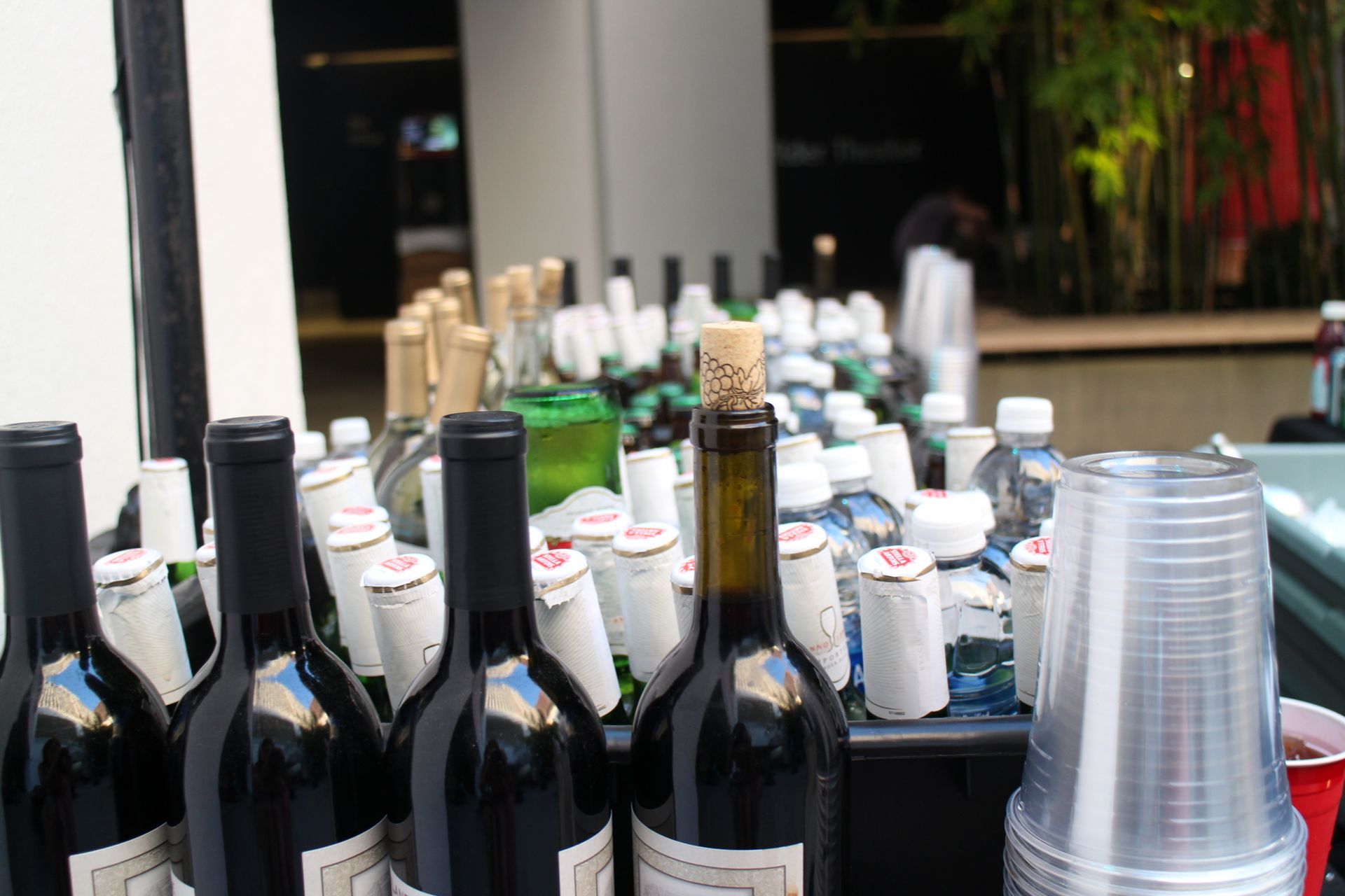 Rows of wine bottles and stacked plastic cups on a table at an outdoor event