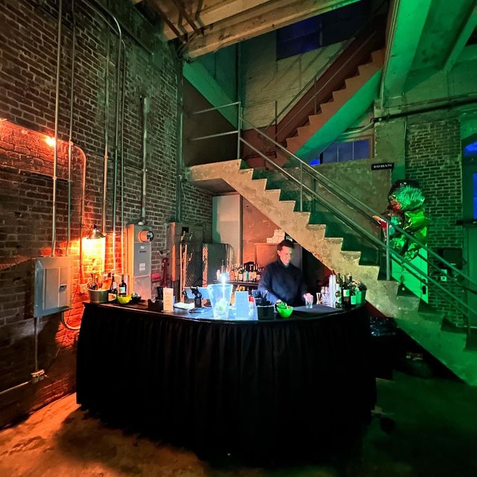 Dimly lit industrial loft with a person behind a black bar under a staircase, lit by green and orange lights