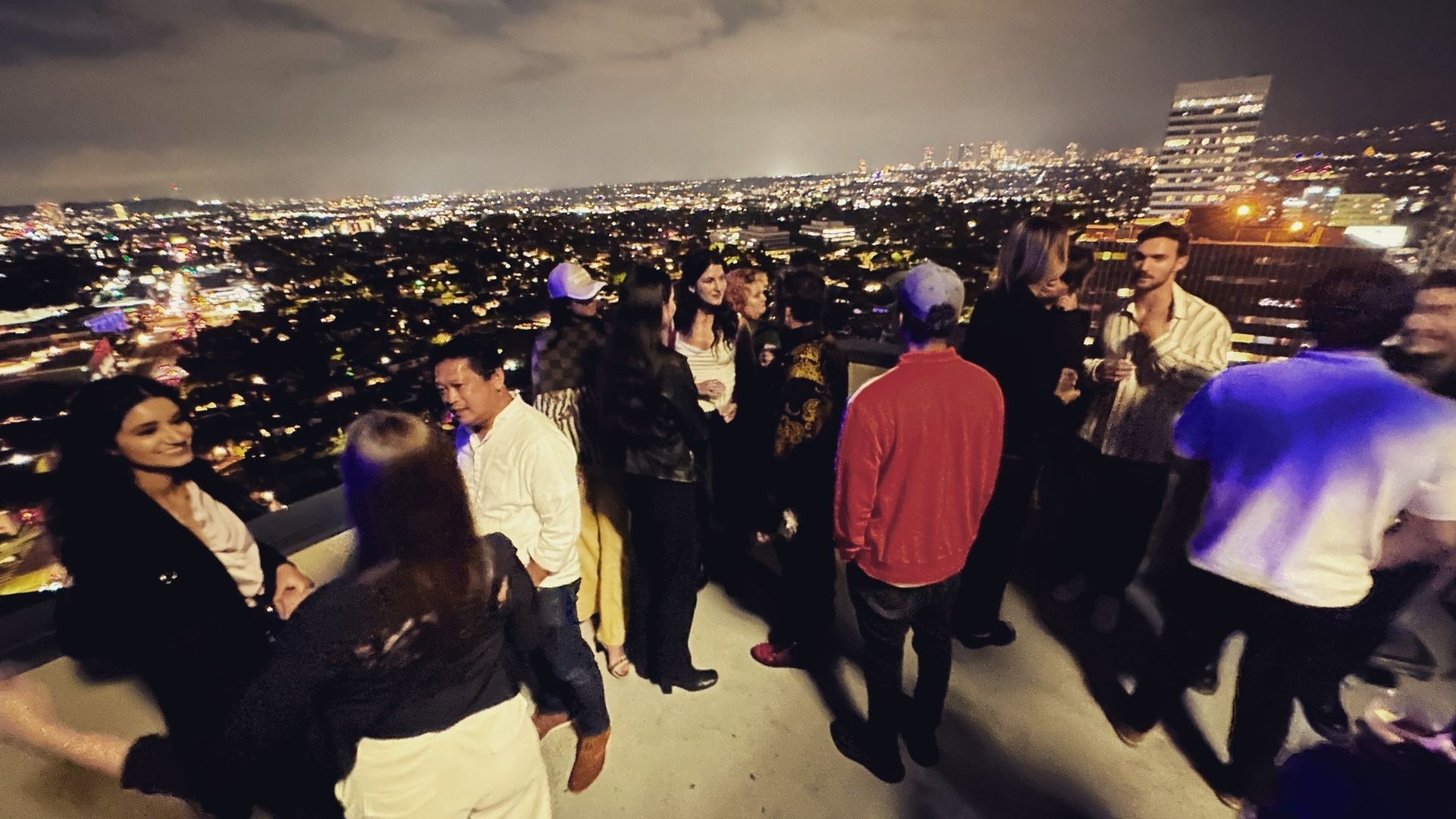 People gathered on a hilltop overlooking a brightly lit city at night.