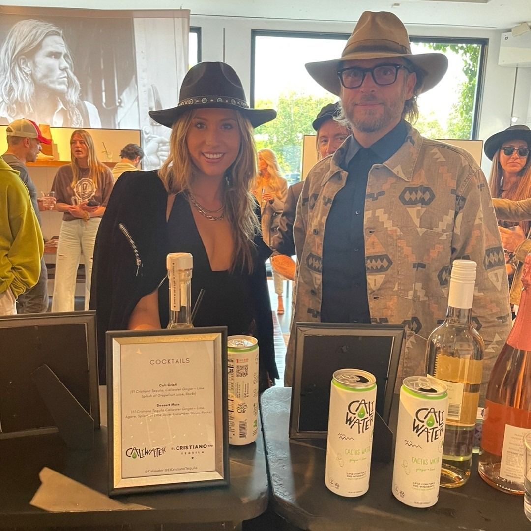 Two people in hats smiling behind a wine tasting table with bottles and labels displayed
