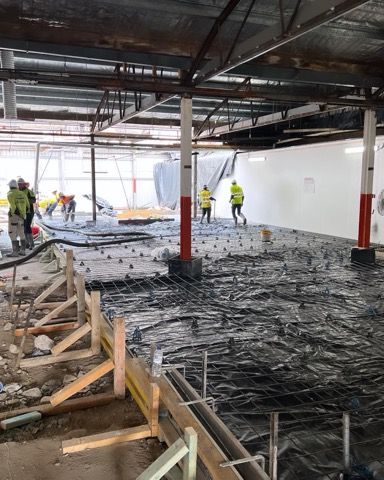 Construction site, workers preparing concrete floor, rebar and formwork visible. White and red support columns.