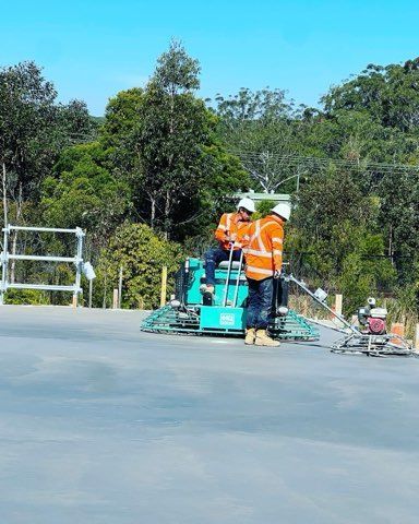 Two construction workers operating a power trowel on a concrete surface, wearing safety vests and helmets.