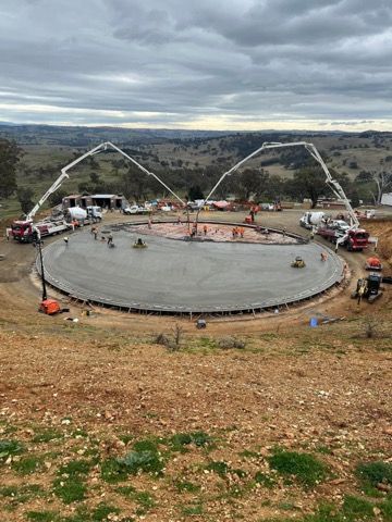 Construction site with circular concrete foundation. Two concrete pump trucks pouring concrete. Overcast sky, rolling hills.