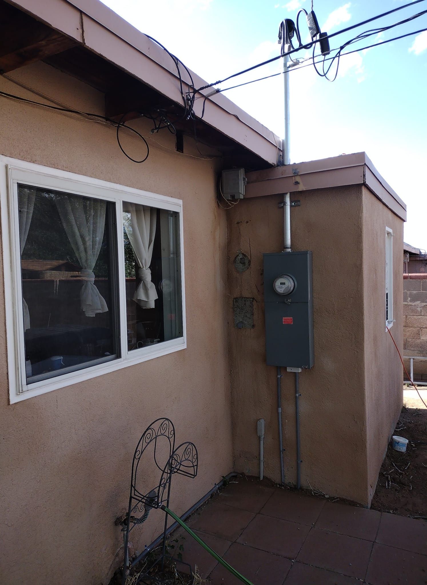 Exterior wall with window, utility meter box, and cables beside a stucco house.