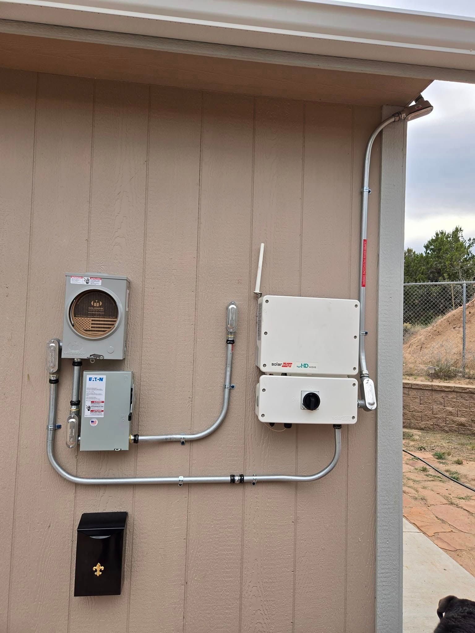 Exterior utility wall with electrical meter, boxes, conduit, and a black wall-mounted panel.