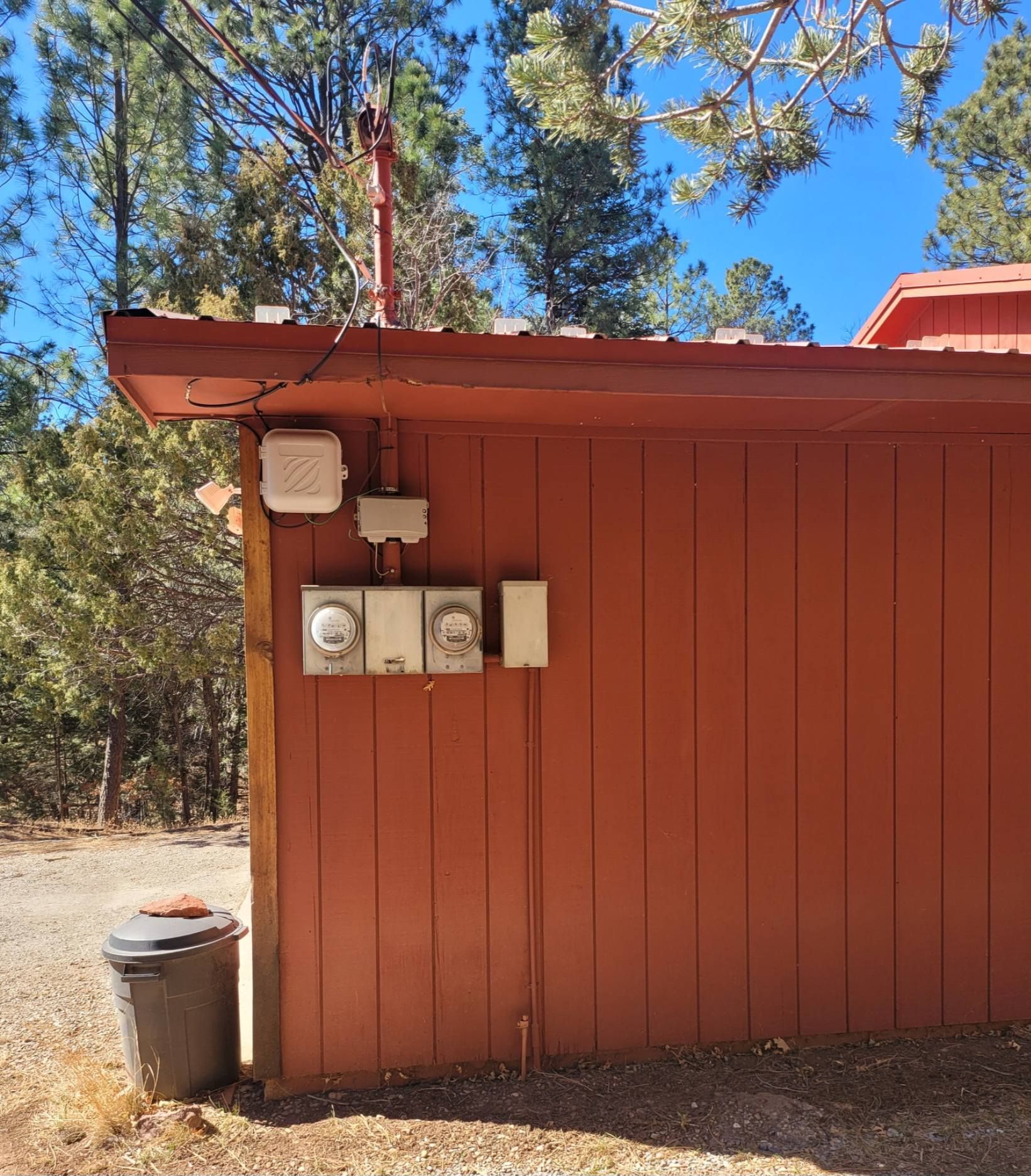 Red utility building wall with meters and cables under a pine tree, with a trash can beside it.