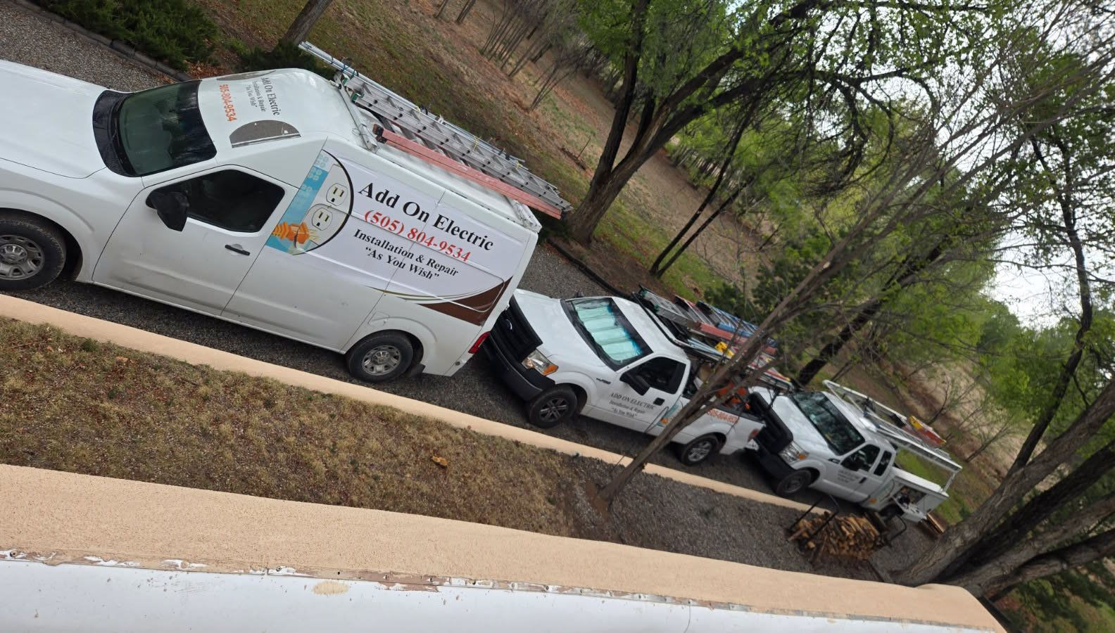 Two white utility vans parked nose-to-tail on a sloped roadside beside trees and grass.