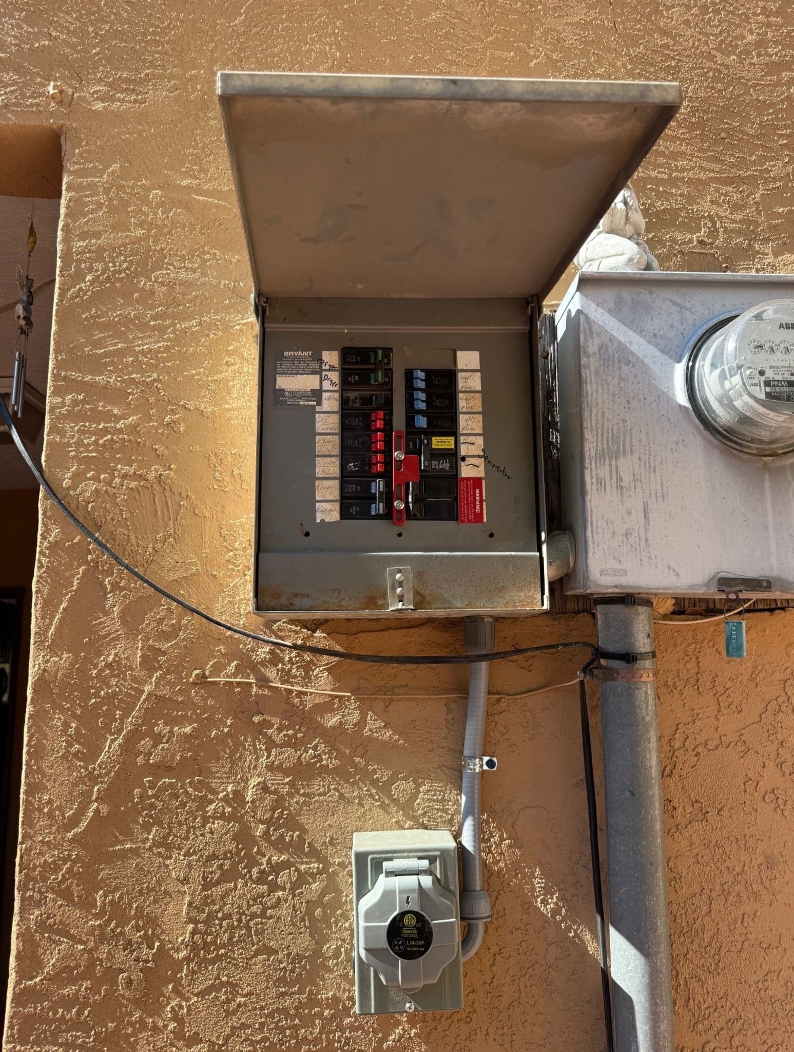 Open electrical breaker box on a tan wall with exposed wiring and a nearby meter.
