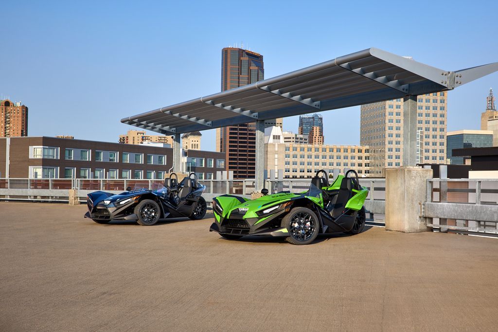 Two slingshot cars are parked under a canopy in a parking lot.