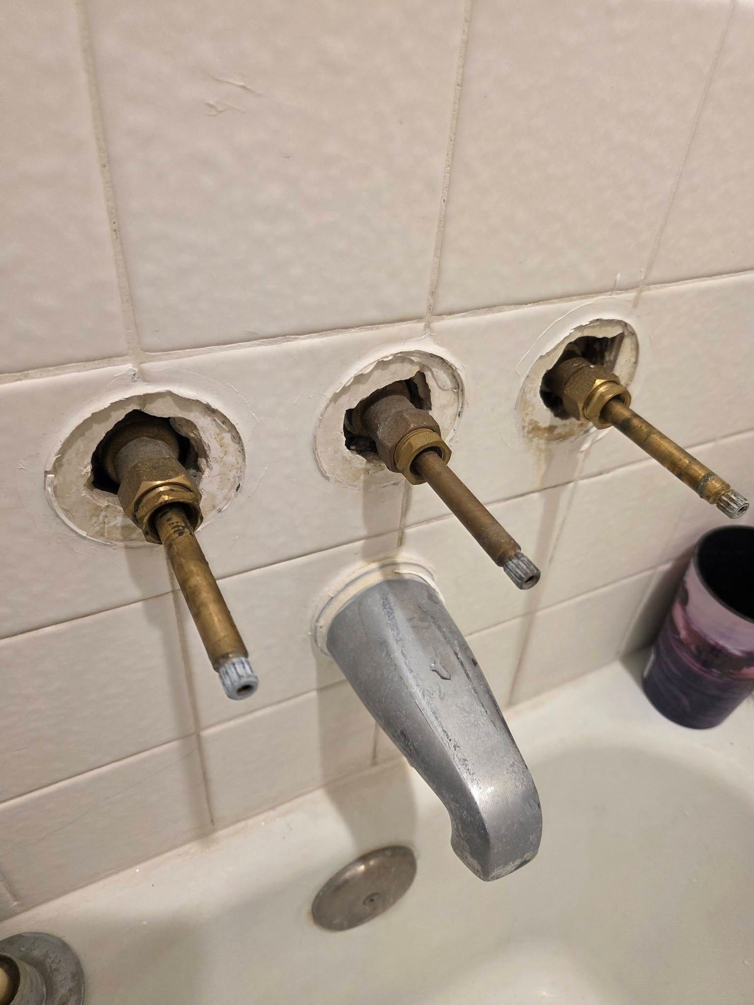 Three exposed faucet valves and a missing sink spout against a beige tiled wall, with a detached handle below.