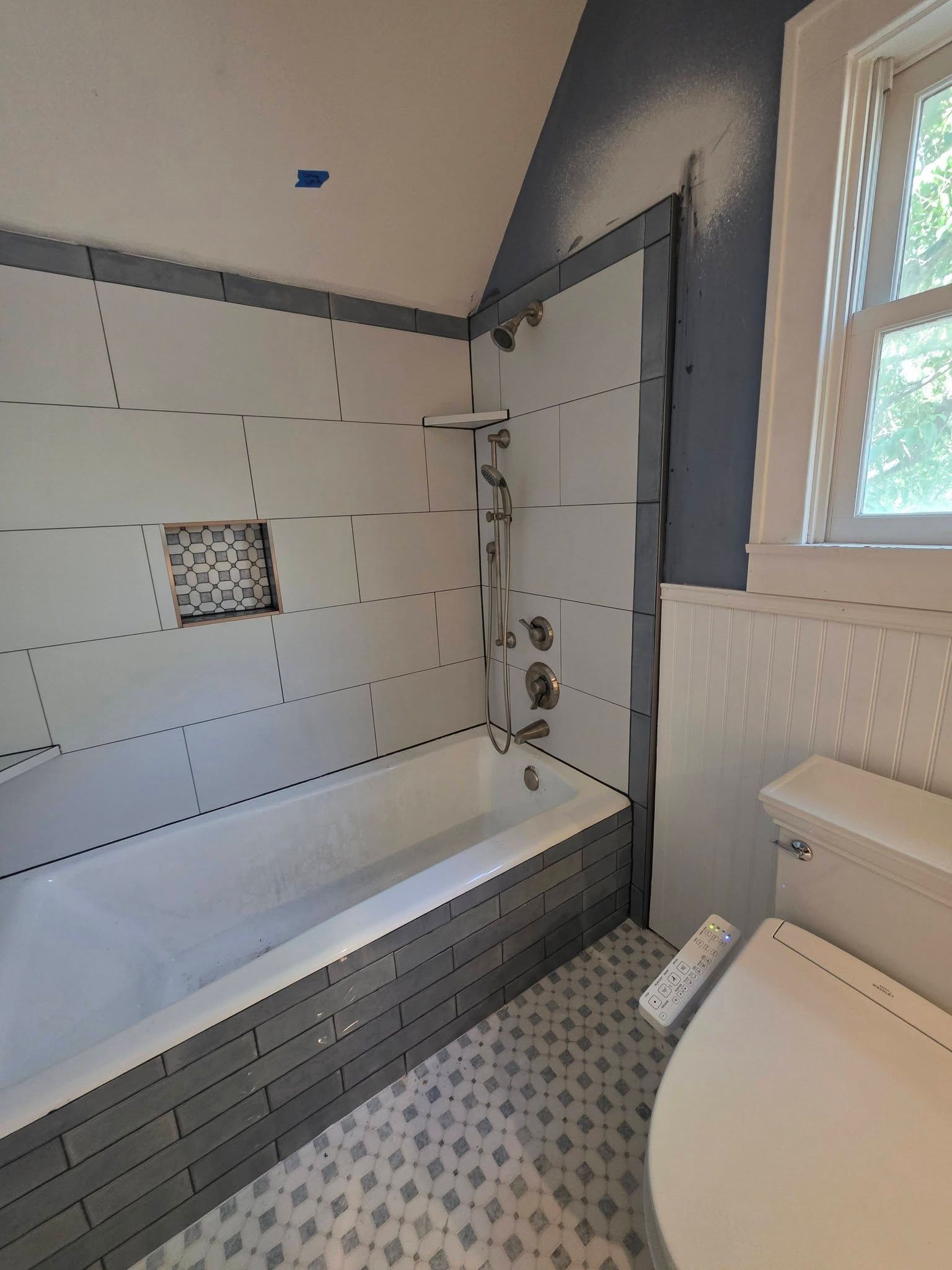 Small bathroom with a white tub-shower, gray tile walls, and a toilet by a window.
