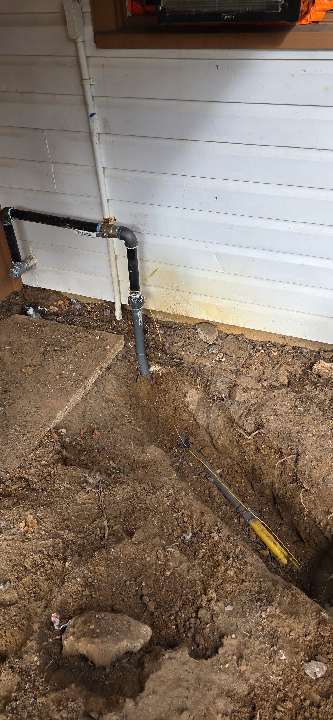 Outdoor dirt area beside a white wall with exposed plumbing pipes and a trench in the ground