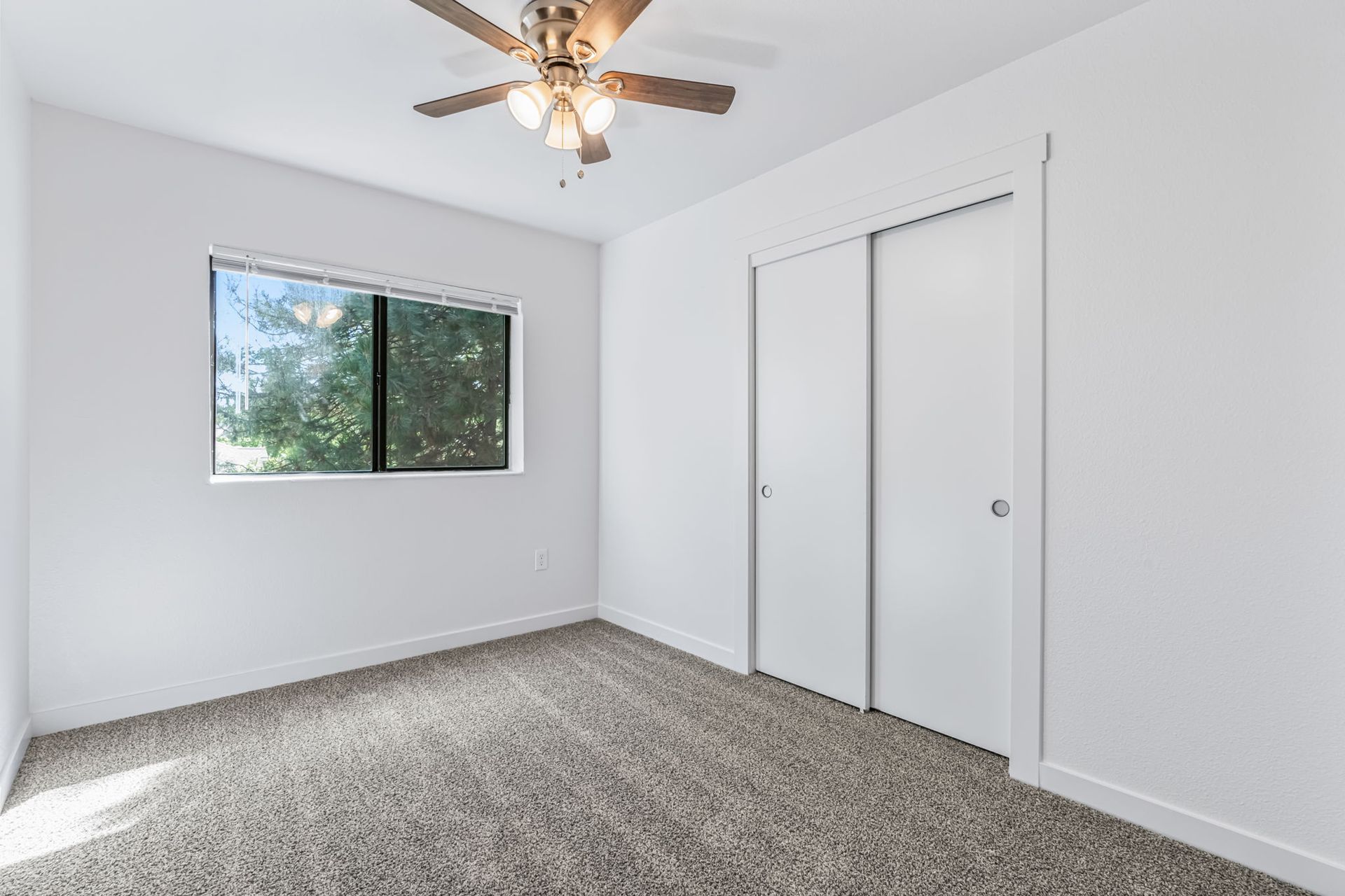 Empty bedroom with neutral carpet, white walls, closet doors, and window with view of trees.