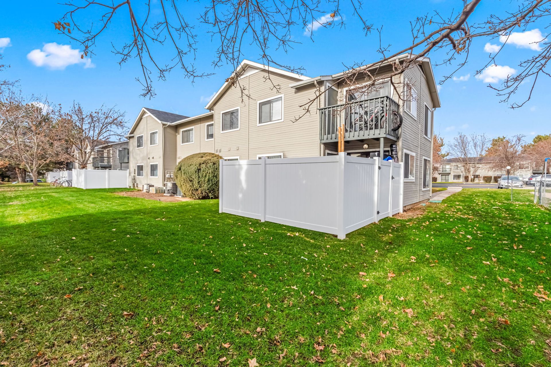 Two-story beige apartments with white fence and green lawn under blue sky.