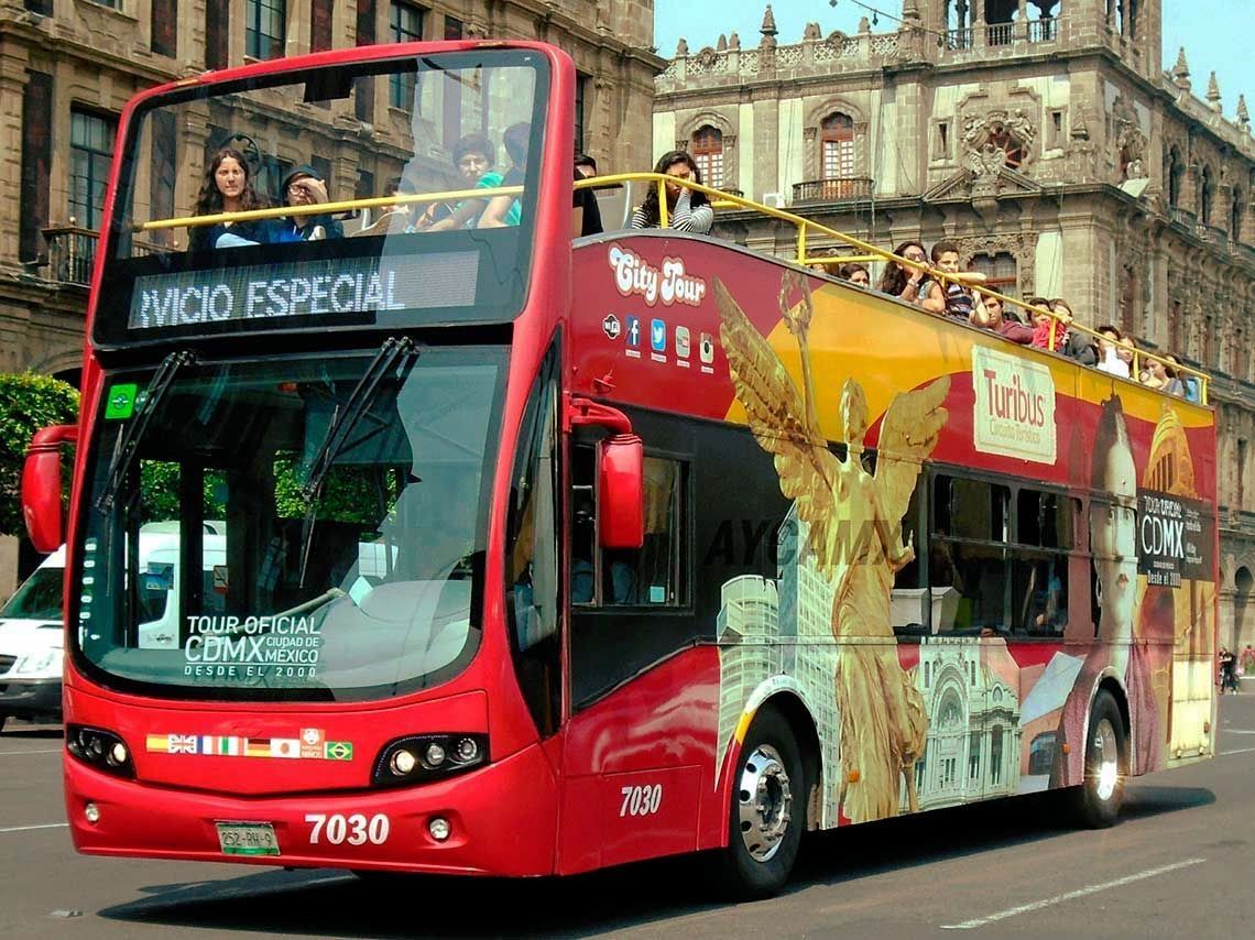 Autobús turístico rojo de dos pisos en la Ciudad de México. Personas en el piso superior, con un edificio al fondo.