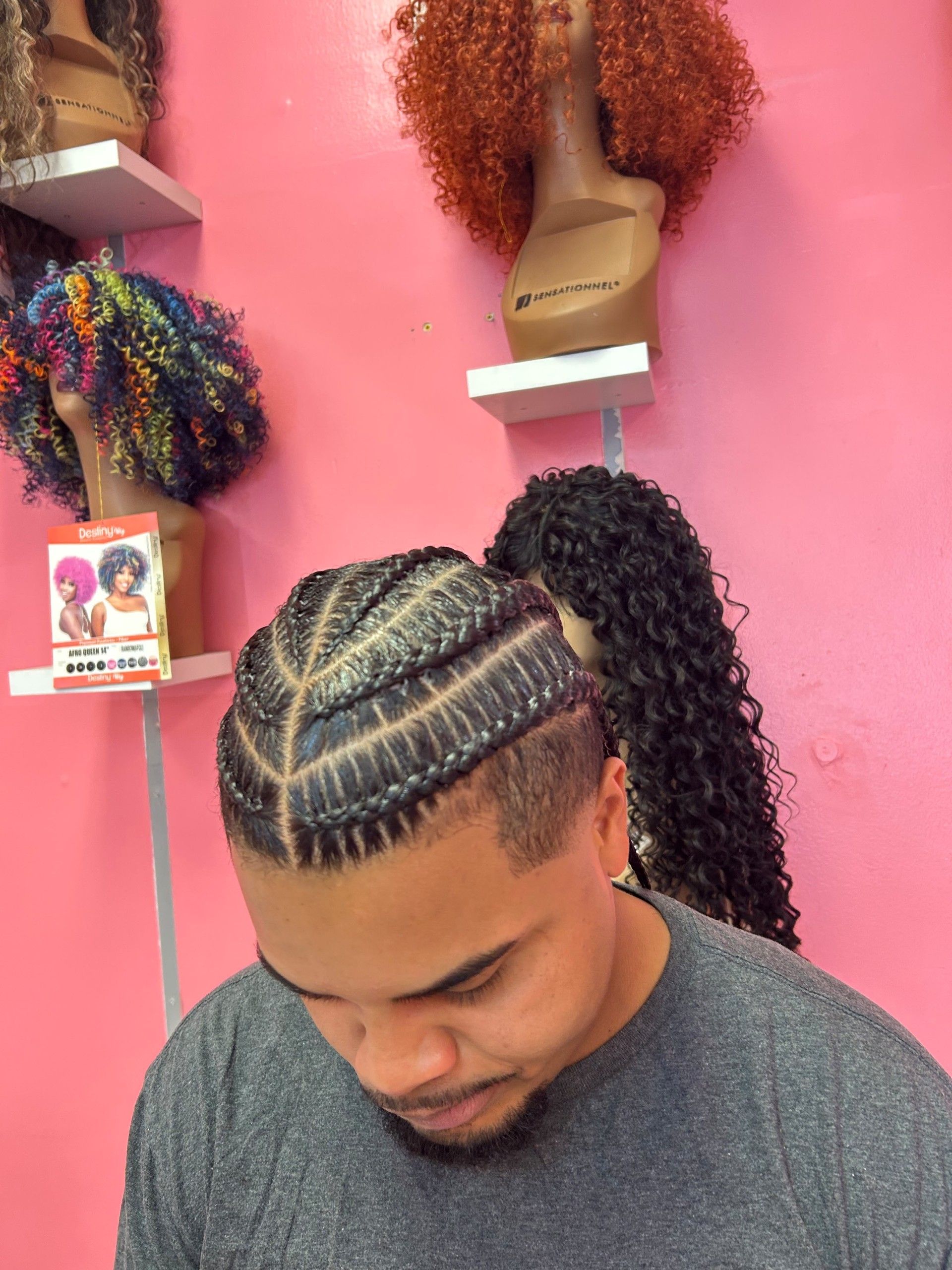 Man with braided cornrows in a pink hair salon, looking down with curly wigs displayed on shelves behind him