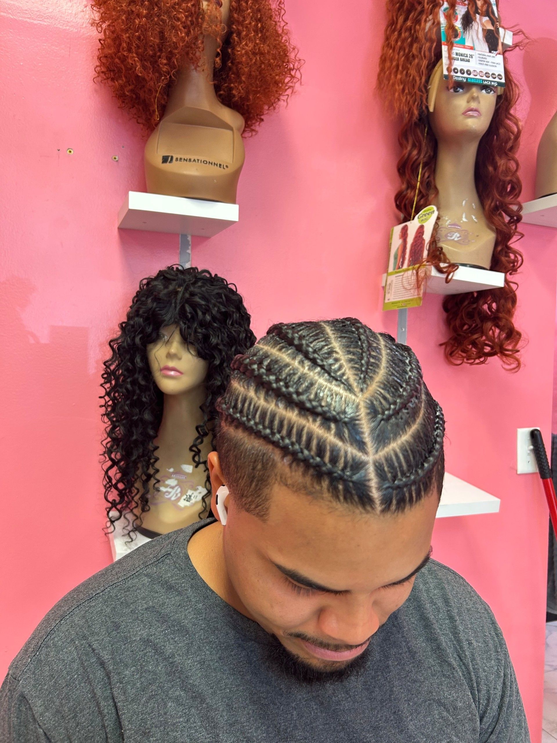 Man with braided hair looking down in a pink wig shop, with mannequin heads and wigs on the wall
