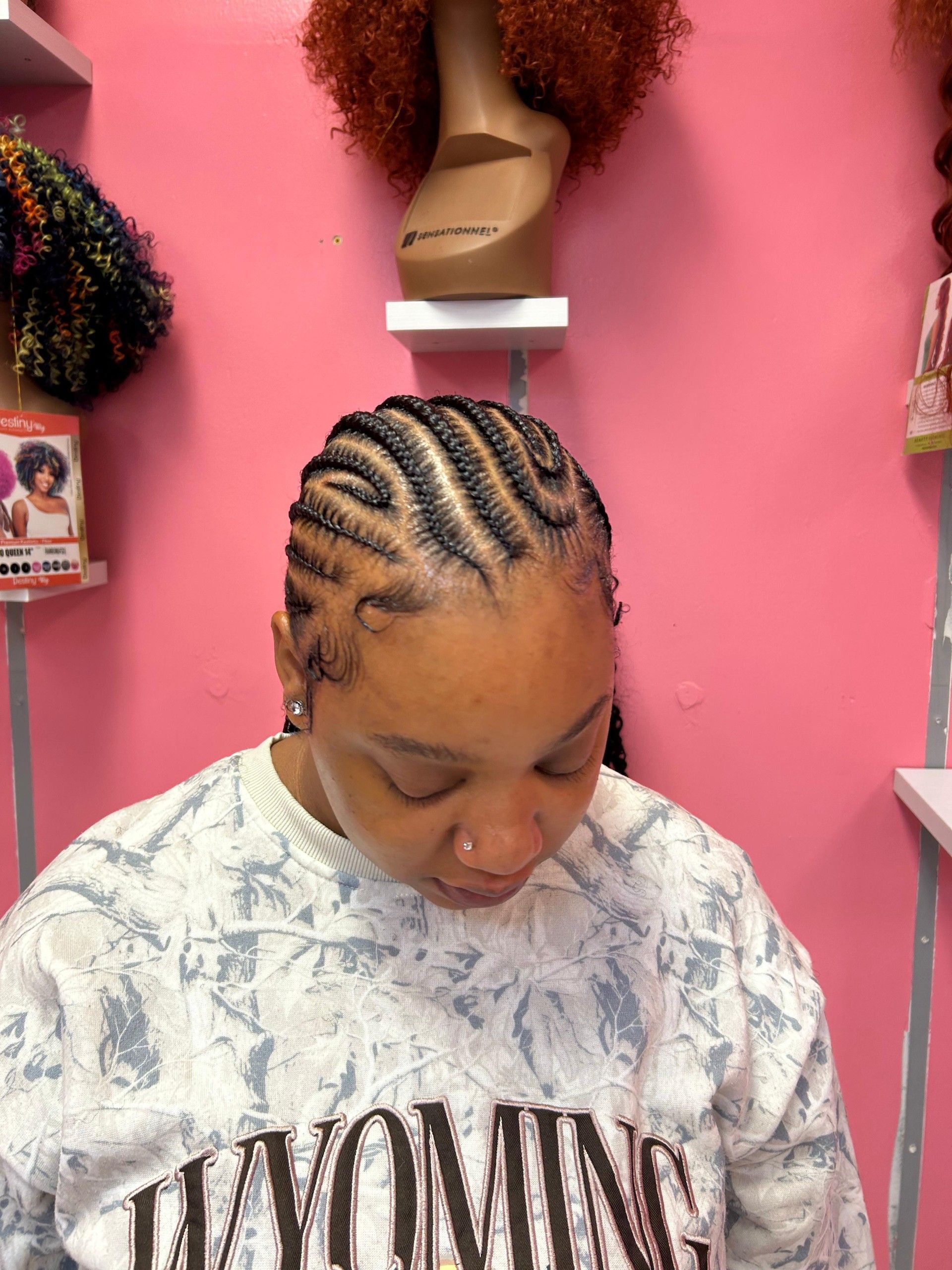 Person with braided hair looking down in a pink salon, wearing a gray Wyoming sweatshirt.